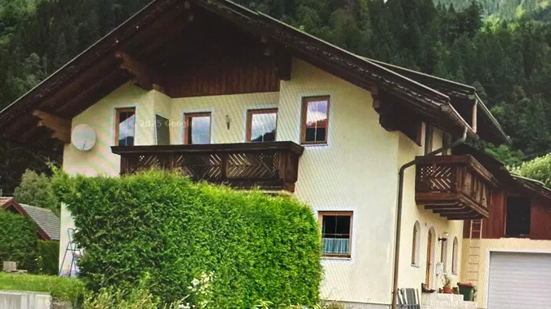 Traditionelles Haus im Alpenstil mit Holzbalkonen und gepflegter Hecke vor einer Bergkulisse.