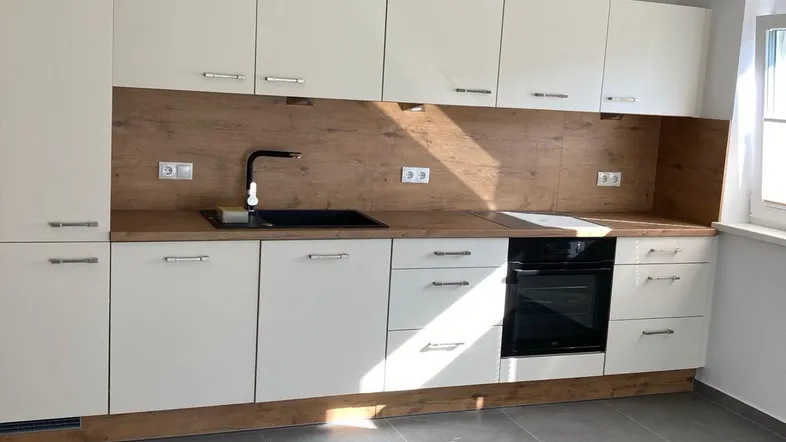 Moderne Küche mit weißen Schränken, Holzdekor und integriertem Backofen, hell durch Tageslicht.