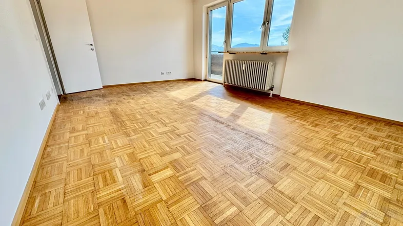 Helles Zimmer mit Parkettboden, großem Fenster und Zugang zum Balkon mit Bergblick.