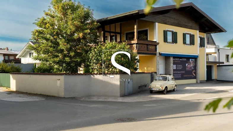 Traditionelles gelbes Gebäude mit Holz-Balkon, grünen Fensterläden und Satteldach in ländlicher Umgebung.