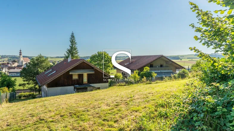 Sonniges Grundstück mit zwei Häusern auf einem Hügel und weitem Blick über die ländliche Umgebung und ein Dorf.