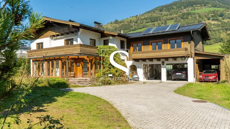 Großzügiges Haus mit Carport, Auffahrt und Solaranlage, eingebettet in eine grüne Berglandschaft.