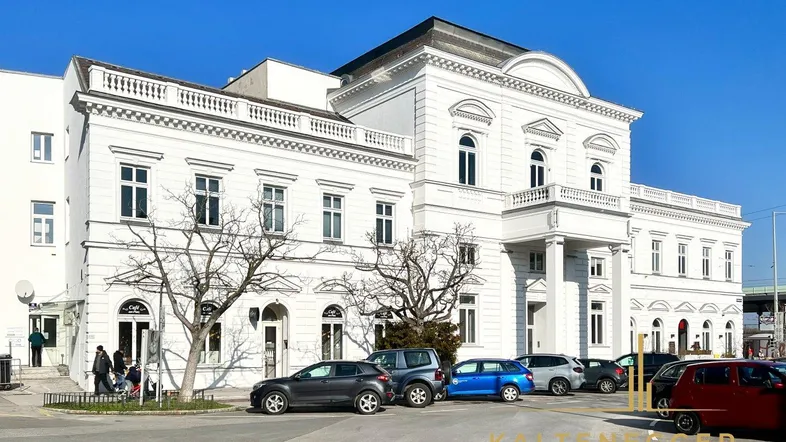 Klassisches weißes Gebäude mit Säulen und Balkon, umgeben von Parkplätzen unter strahlend blauem Himmel.