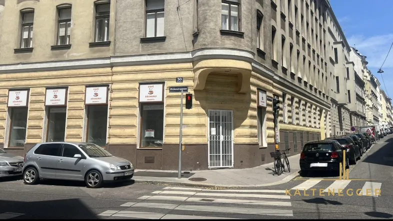 Gebäudeecke an einer belebten Straße mit parkenden Autos und Fußgängerüberweg in städtischer Umgebung.
