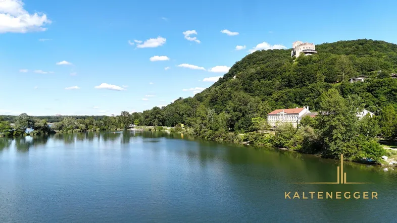 Weitläufiger Blick auf einen ruhigen See mit bewaldeten Hügeln und einem historischen Gebäude in der Ferne.