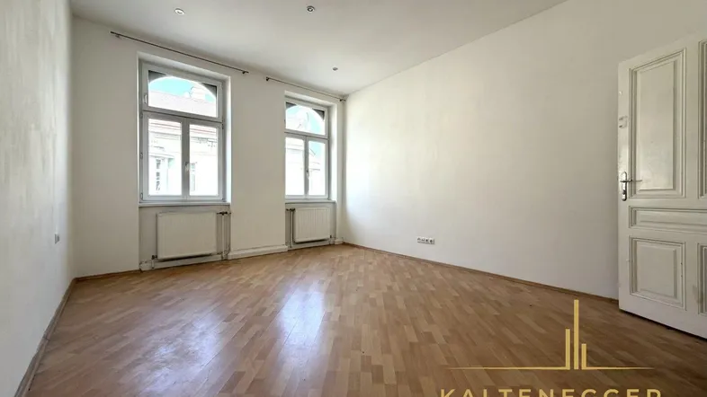 Geräumiges, leeres Zimmer mit Parkettboden, hohen Decken und einer klassischen Holztür.