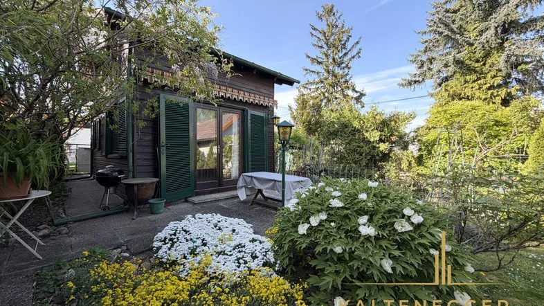 Gemütlicher Gartenbereich mit blühenden Pflanzen, Sitzgelegenheit und einem charmanten Holzhaus im Hintergrund.