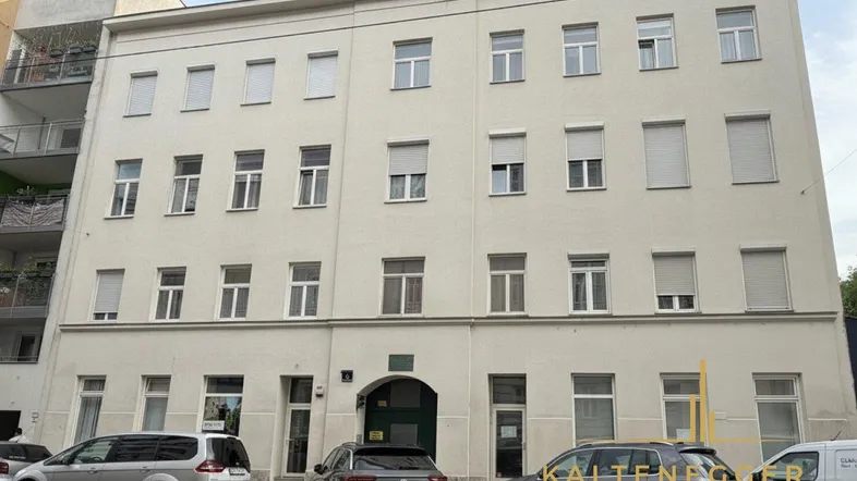 Fassade eines Mehrfamilienhauses mit mehreren Fenstern und geparkten Autos davor in einer städtischen Umgebung.