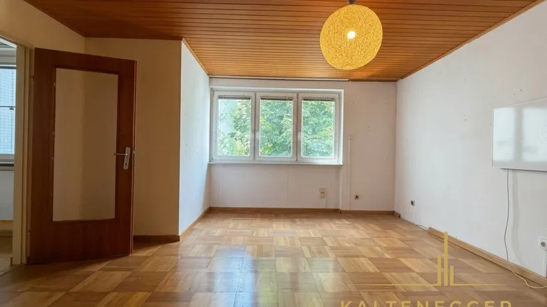 Geräumiges Zimmer mit Fischgrätparkett, Holzdecke und großen Fenstern, die viel Tageslicht hereinlassen.