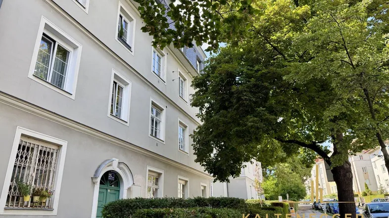 Klassisches Mehrfamilienhaus mit heller Fassade und grüner Hecke entlang des Bürgersteigs.