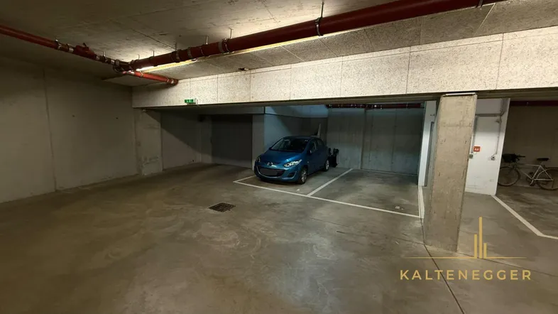 Geräumige Tiefgarage mit Betonwänden und -boden, einem geparkten blauen Auto und sichtbaren roten Rohren.