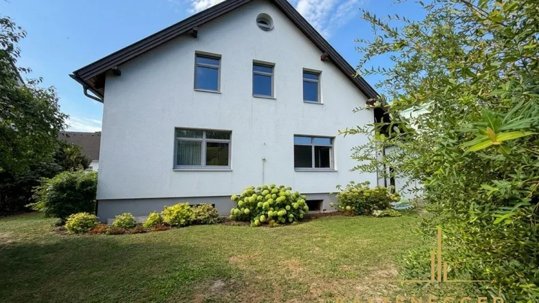 Ansicht des weißen Einfamilienhauses mit gepflegtem Vorgarten und blühenden Sträuchern unter blauem Himmel.