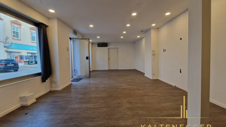 Geräumiger, leerer Geschäftsraum mit großen Schaufenstern, modernem Laminatboden und Einbaustrahlern an der Decke.