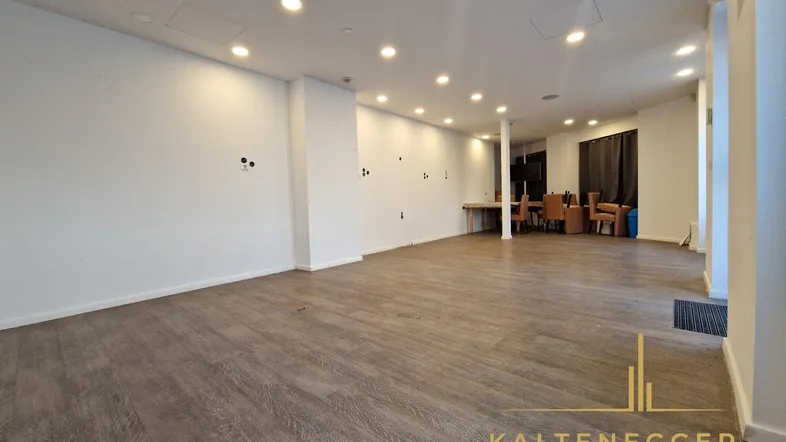 Großzügiger, leerer Verkaufsraum mit hellem Laminatboden, weißen Wänden und moderner Deckenbeleuchtung, bereit zur Nutzung.