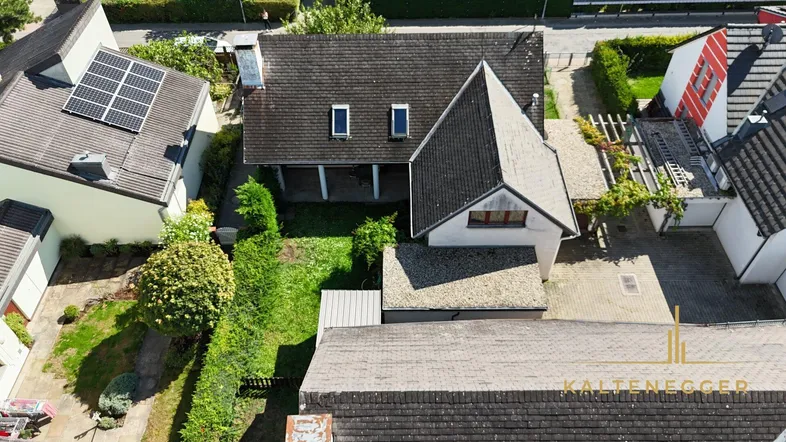 Luftaufnahme eines Einfamilienhauses mit gepflegtem Garten und angrenzenden Gebäuden unter blauem Himmel.
