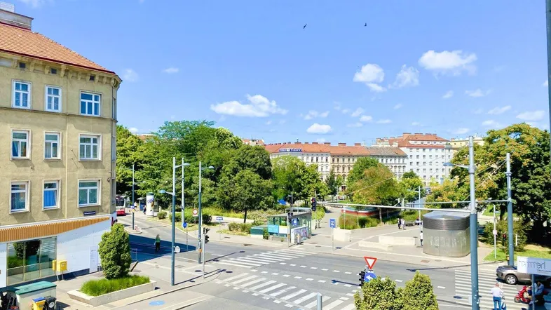 Blick auf eine belebte Stadtkreuzung mit Gebäuden, Bäumen und blauem Himmel an einem sonnigen Tag.