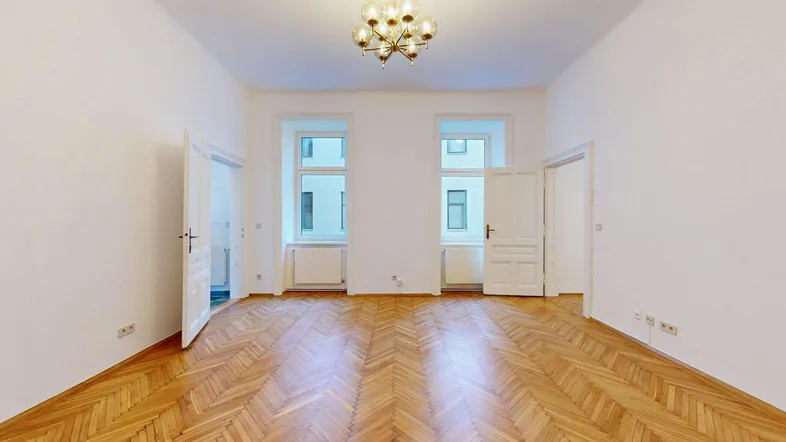 Geräumiges Wohnzimmer mit Fischgrätparkett, hohen Decken und zwei großen Fenstern, die viel Tageslicht hereinlassen.