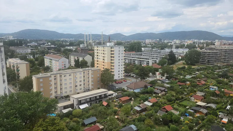 Weitläufiger Blick über die Stadt mit Wohngebäuden, Schrebergärten und Bergen im Hintergrund.