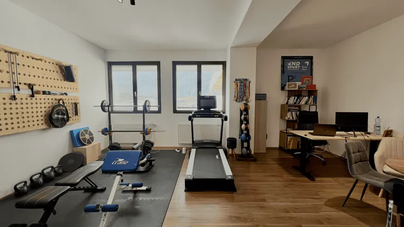 Großer Raum mit Fitnessbereich und Arbeitsplatz, ausgestattet mit Laufband, Gewichten und Schreibtischen.