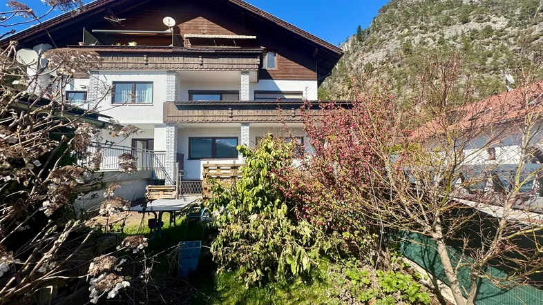 Mehrstöckiges Wohnhaus mit Gartenbereich und Balkonen, umgeben von grüner Natur und Bergen.