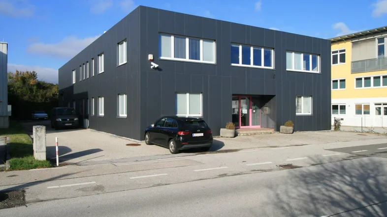 Modernes Geschäftsgebäude mit dunkler Fassade und hellen Fenstern, umgeben von einem Parkplatz.