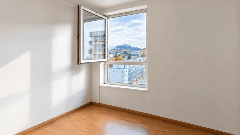 Helles Zimmer mit Holzboden und einem Fenster, das einen Blick auf die Stadt und Hügel bietet.