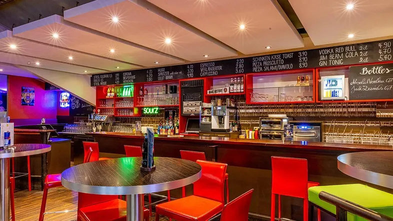 Moderne Bar mit leuchtend roten Stühlen, einer umfangreichen Getränkekarte und Deckenbeleuchtung.