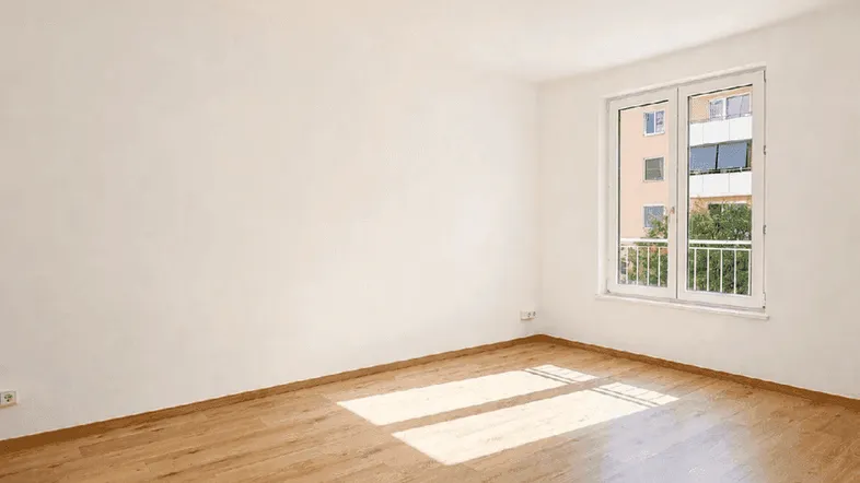Helles Zimmer mit Holzfußboden und Fenster, das viel Tageslicht hereinlässt und einen kleinen Balkon zeigt.