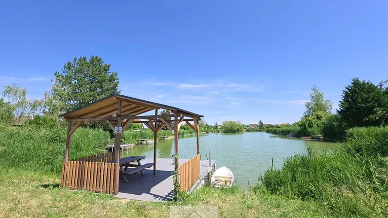 Holzpavillon mit Picknicktisch am Ufer eines ruhigen Sees unter blauem Himmel.