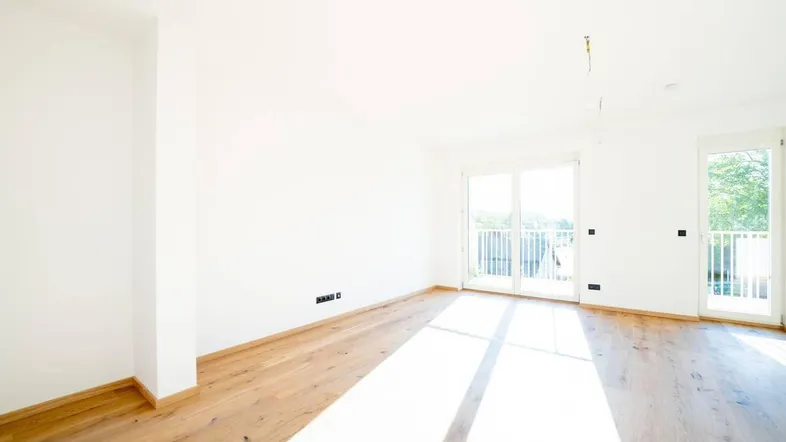 Helles, leeres Wohnzimmer mit Holzboden und Zugang zu einem Balkon, durchflutet von Sonnenlicht.