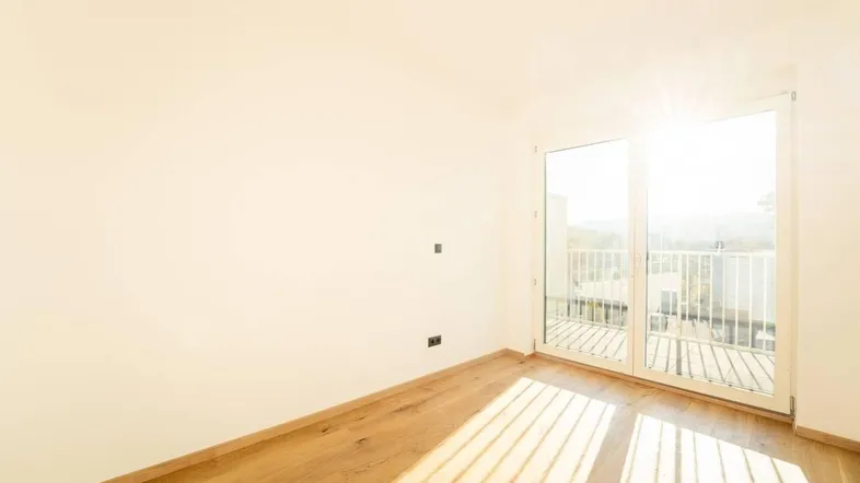 Heller, leerer Raum mit Holzboden und Zugang zu einem sonnigen Balkon.