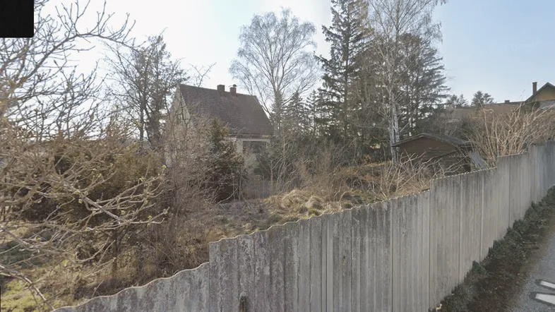 Blick auf ein Grundstück mit älterem Haus, umgeben von Bäumen und einem Holzzaun, aufgenommen von der Straße.