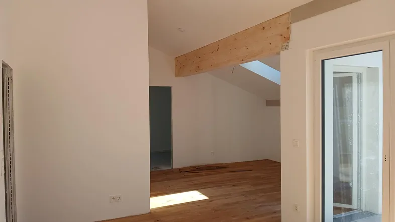 Heller Wohnbereich mit Holzboden, sichtbarem Holzbalken, Dachschräge und Zugang zu einem Außenbereich.