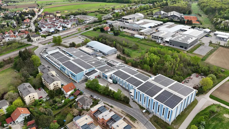 Luftaufnahme eines großen Industriekomplexes mit Solaranlagen auf den Dächern, umgeben von Wohngebäuden und Feldern.