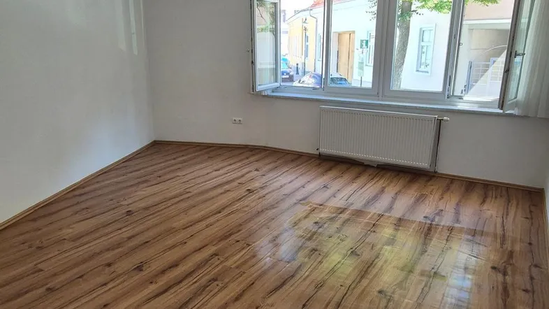 Helles Zimmer mit Laminatboden und großen Fenstern, die viel Tageslicht hereinlassen.