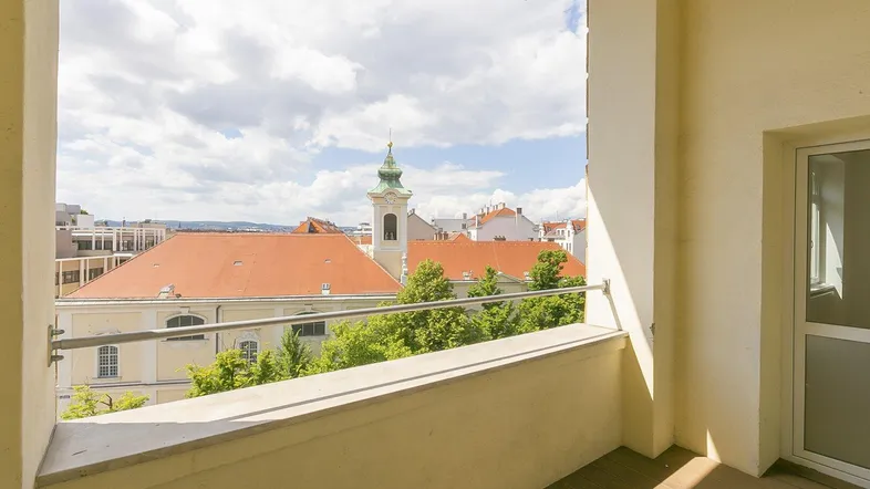 Heller Balkon mit Holzboden und Blick auf die Stadt und eine Kirche unter blauem Himmel.