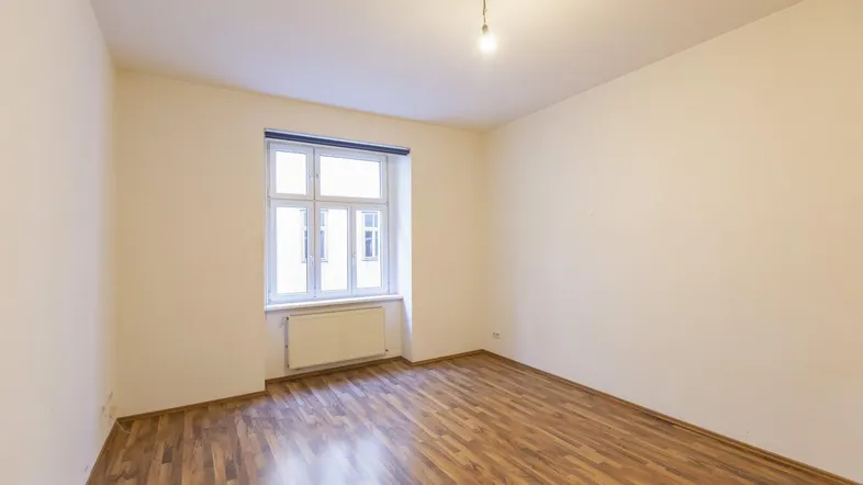 Helles, leeres Zimmer mit Laminatboden und einem Fenster, das viel Tageslicht hereinlässt.