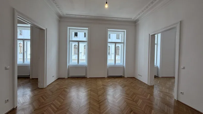 Helles Zimmer mit Fischgrätparkett, hohen Decken und Stuckverzierungen, ideal als Wohnbereich.