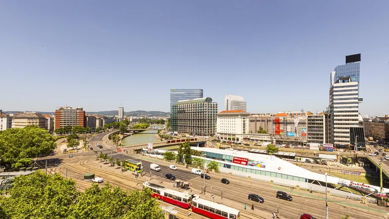 Weitläufige Stadtansicht mit Straßenbahnen, Fluss und modernen Gebäuden unter klarem Himmel.