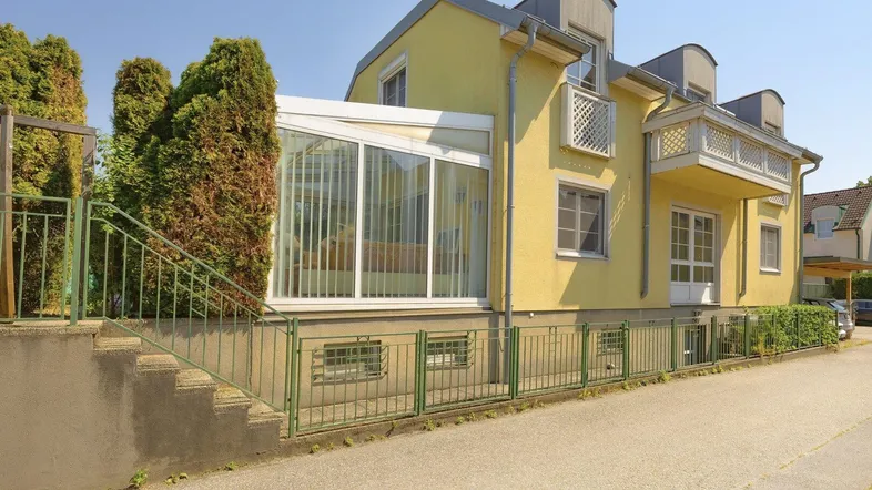 Gelbe Hausfassade mit Wintergarten und Außentreppe, umgeben von grünen Büschen und einem Zaun.
