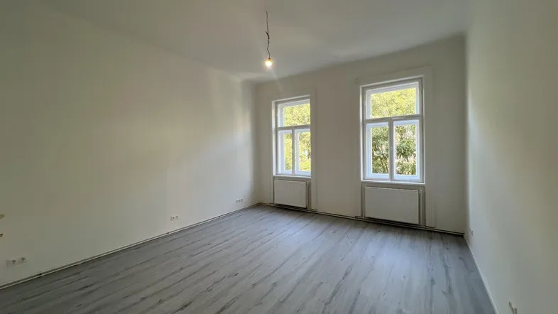 Helles, leeres Zimmer mit grauem Laminatboden und zwei Fenstern, die viel Tageslicht hereinlassen.