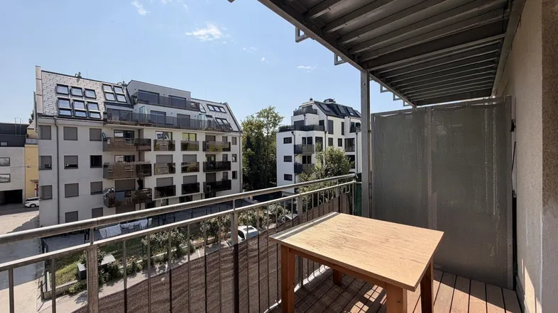Balkon mit Holzboden und Blick auf umliegende moderne Wohngebäude unter blauem Himmel.