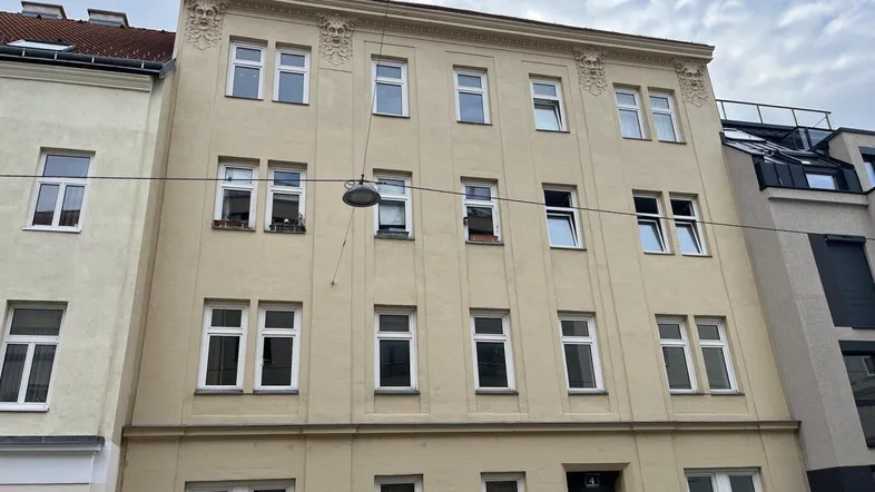 Fassade eines gepflegten Mehrfamilienhauses mit beiger Putzfassade und dekorativen Elementen am Dachrand.