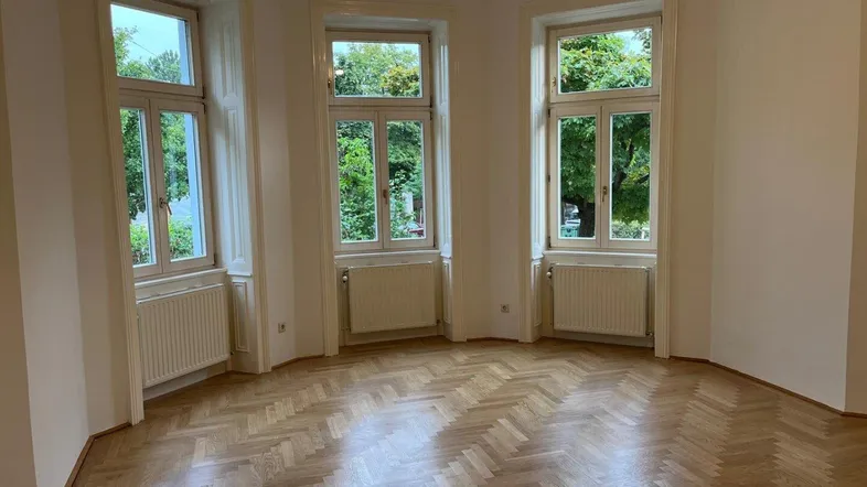 Helles Wohnzimmer mit Fischgrätparkett und drei großen Fenstern mit Blick ins Grüne.
