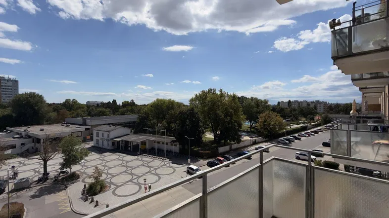 Blick vom Balkon auf die Straße, umliegende Gebäude und grüne Bäume unter blauem Himmel.