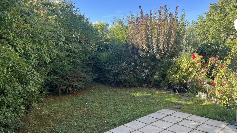 Gepflegter Gartenbereich mit Rasenfläche und gepflasterter Terrasse, umgeben von üppigem Grün.