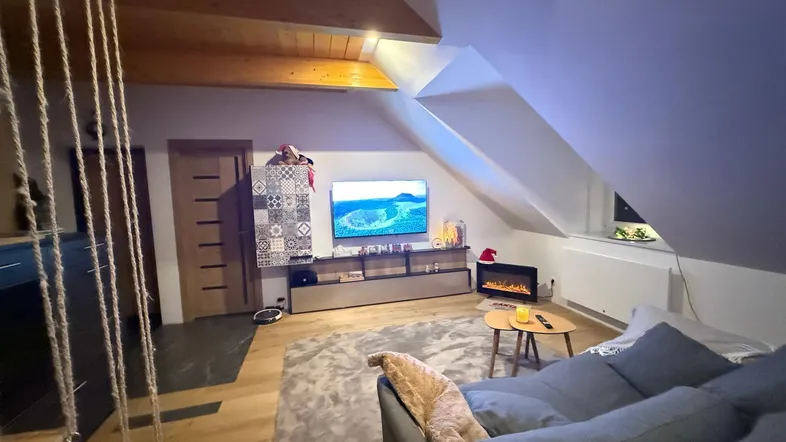 Gemütliches Dachgeschoss-Wohnzimmer mit Holzbalken, Fernseher und dekorativem Elektrokamin.
