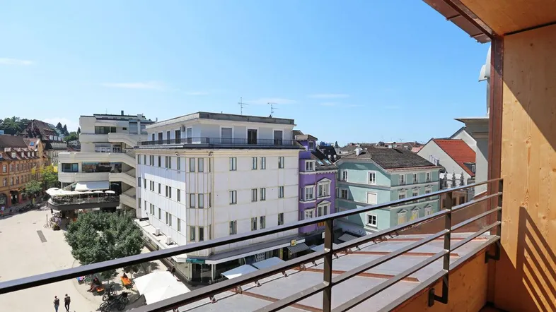 Blick vom Balkon auf den belebten Stadtplatz mit umliegenden Gebäuden und blauem Himmel.
