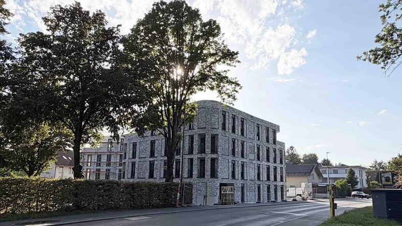 Modernes Mehrfamilienhaus mit grauer Ziegelfassade und großen Fenstern, umgeben von Bäumen.