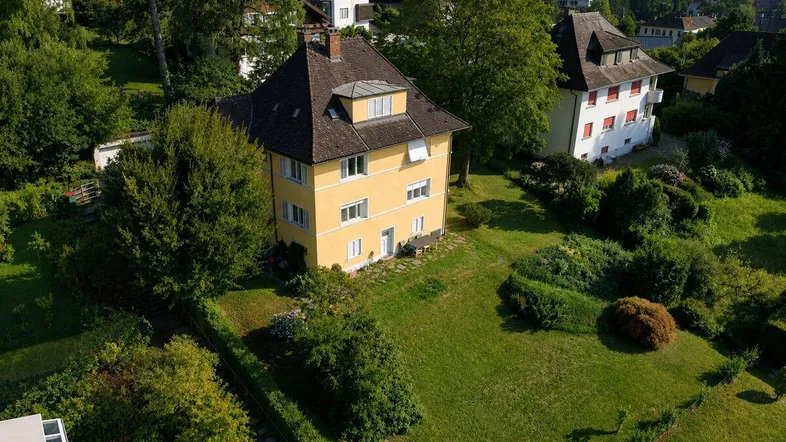 Luftaufnahme eines gelben Hauses mit großem Garten und umliegenden Bäumen in sonniger Lage.